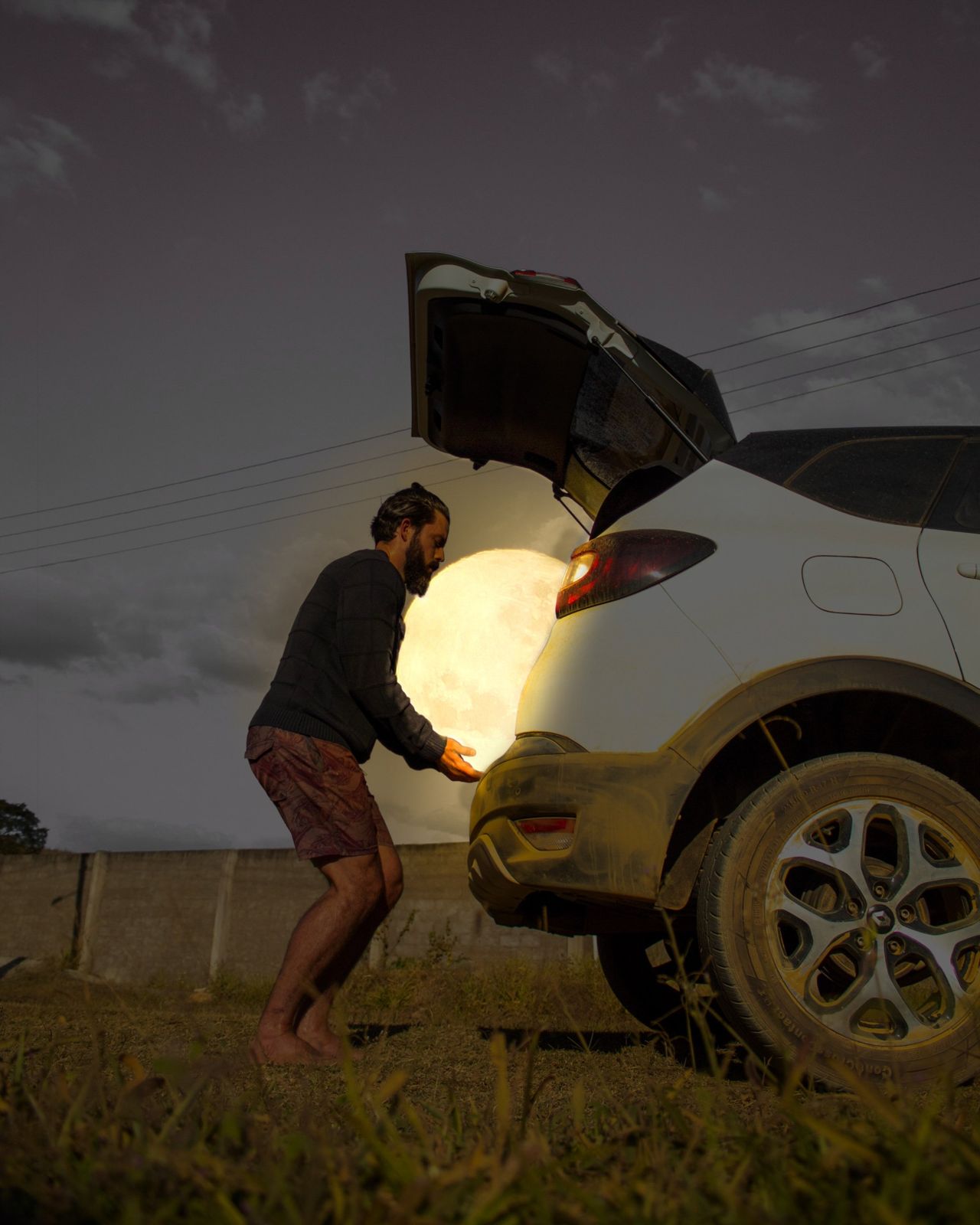 Man loading the full Moon into the open trunk of a white car at dusk, surreal photograph by Daniel Antoniol